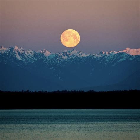 The Full Moon Is Setting Over Mountains In The Distance With Water And
