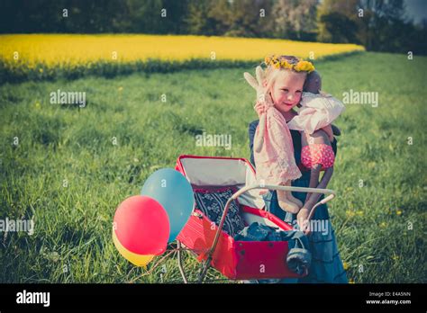 Smiling Little Girl Holding Doll And Soft Toy Standing On Meadow With
