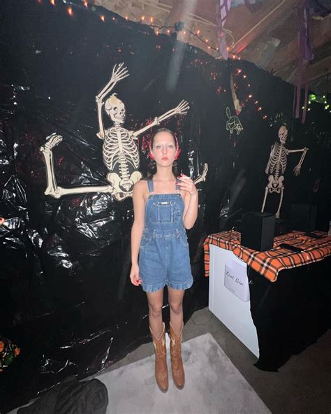 A Woman Standing In Front Of Skeletons On Display At A Halloween Event With Her Phone Up To Her Ear