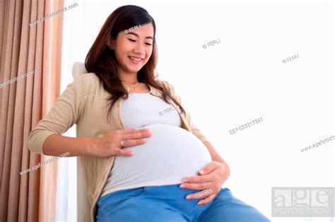 a portrait of a happy pregnant asian woman touching her big belly while sitting on a chair