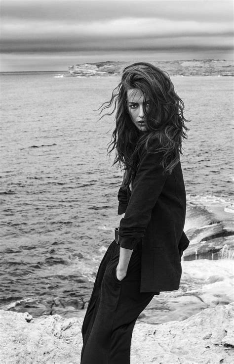 A Black And White Photo Of A Woman Standing On Rocks By The Water With Her Hair Blowing In The Wind