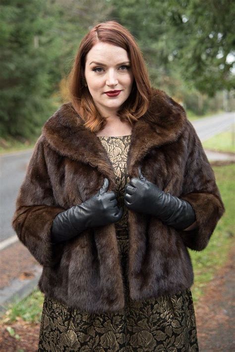 A Woman In A Fur Coat And Black Gloves Standing On The Side Of A Road