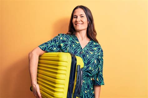Young Beautiful Brunette Tourist Woman Holding Airplane Cabin Suitcase Over Yellow Background