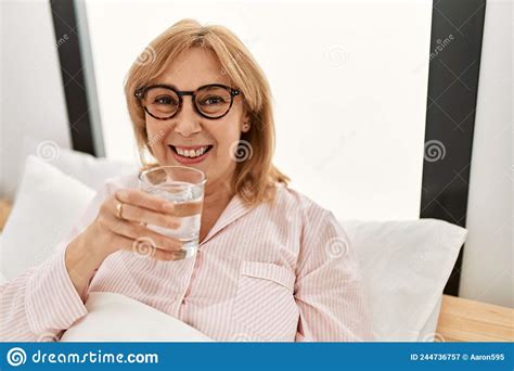Middle Age Blonde Woman Smiling Happy Drinking Glass Of Water Lying On The Bed At Home Stock