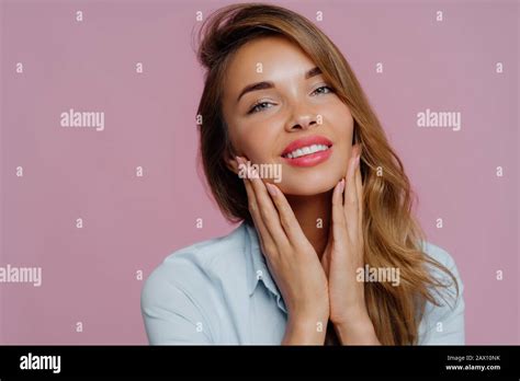 Photo Of Lovely Young Female Model Touches Gently Chin With Both Hands