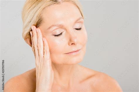 Attractive And Blonde Mature Woman With Closed Eyes Touching Her Face Isolated On Grey Stock