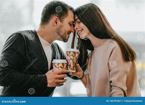 Loving Couple Is Siting Face To Face On The Background Of Window And Holding Cups In Their Hands