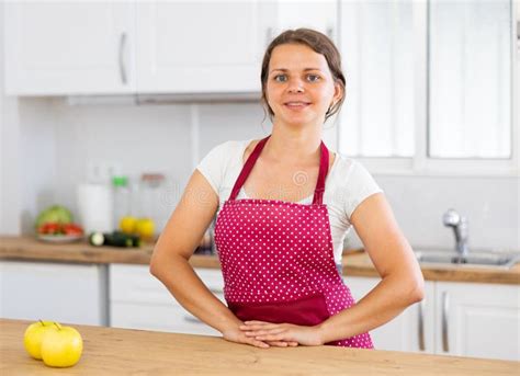 Portrait Of Positive Girl Housewife In Apron Standing At Kitchen Stock