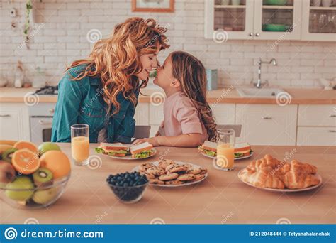 Side View Of Babe And Mother Touching Noses While Sitting At The Kitchen Table With Food