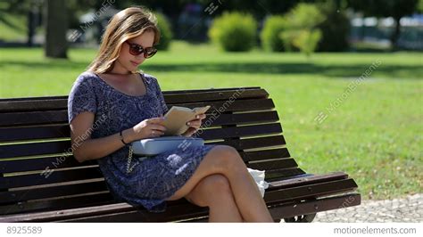 Young Woman Reading A Book And Sitting On A Bench Outside In A Park In