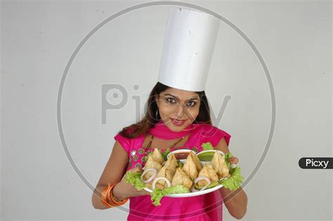 Image Of An Indian Woman Chef In Kitchen Apron And Cap Holding Samosas Plate With An Expression
