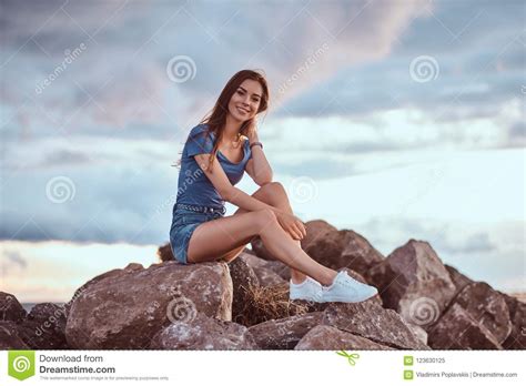 Portrait Of A Girl Sitting On Rocks Against The Cloudy Sky During