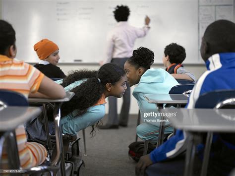 Two Girls Talking In Class While Teacher Is Writing On Board Foto De