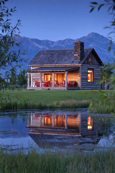 A Small Log Cabin Sits On The Edge Of A Pond At Dusk With Mountains In