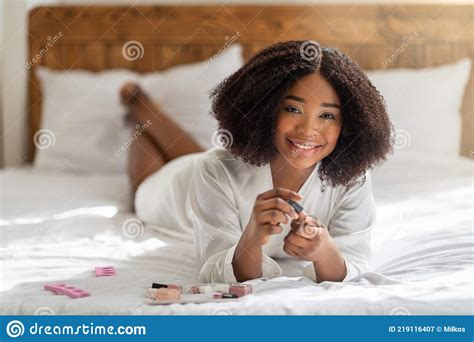 Happy Black Woman Lying On Bed In Dressing Gown Applying Nail Polish