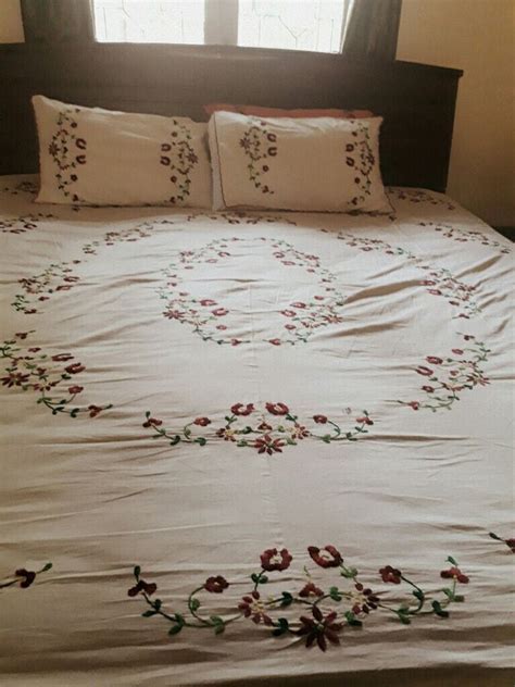 A White Bed With Red And Green Flowers On The Comforter Is In Front Of A Window