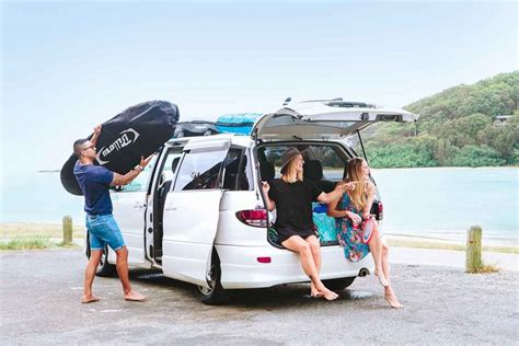 Three People Sitting On The Back Of A Van With Their Surfboards