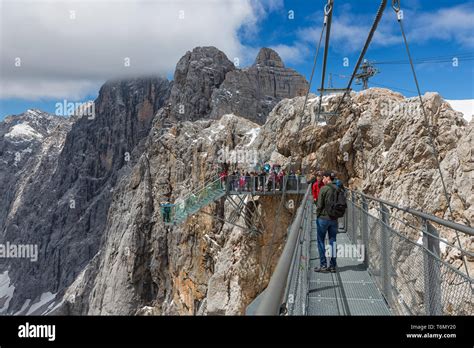Austrian Dachstein Mountains With Hikers Passing A Steel Rope Bridge