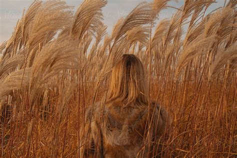 A Blond Haired Girl In A Red Fur Coat Stands In Tall Grass Turning Away From The Camera By