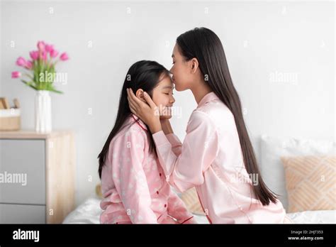Cheerful Japanese Millennial Female Kissing At Forehead Of Teenage Girl In Bedroom Interior
