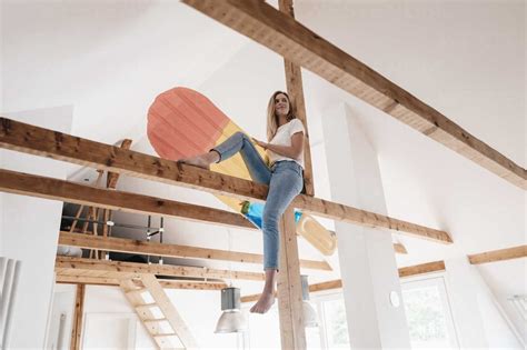 Carefree Young Woman Sitting On Ceiling Joist Holding Colorful Airbed