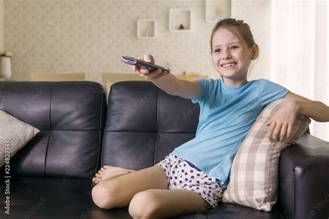 Cute Preteen Girl Watching Tv On Couch Using Remote Control Living Room Interior In Background