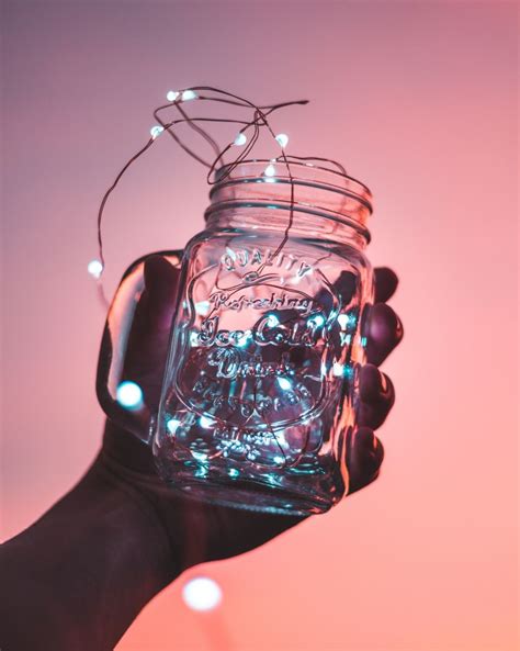 A Hand Holding A Mason Jar With Fairy Lights On It In Front Of A Pink Sky