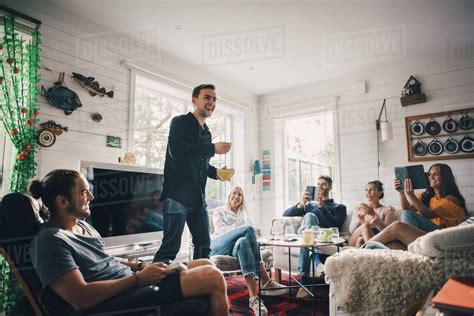 Cheerful Man Standing Amidst Young Friends Sitting In Living Room At