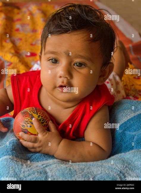 Cute Indian Baby Playing With A Ball Lying On A Yellow Printed Bed