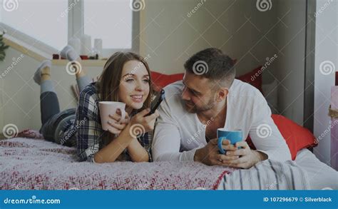 Усмехаясь пары смотря ТВ пока лежащ в кровати и выпивая кофе дома в утре Стоковое Изображение