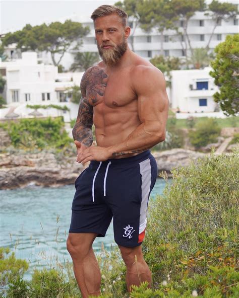 A Man With A Beard And No Shirt Is Standing In The Grass Near Water Wearing Shorts