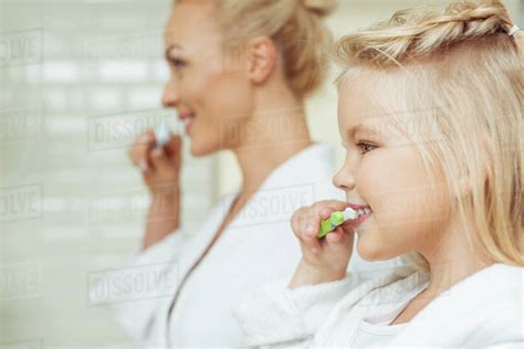 Side View Of Happy Mother And Daughter In Bathrobes Brushing Teeth