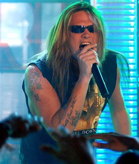 A Man With Long Hair And Sunglasses Singing Into A Microphone In Front