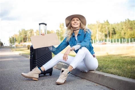 Smiling Solitude Blond Woman In Hat Hitchhiking Sit On Curb Near Grass Hold Blank Carton Board