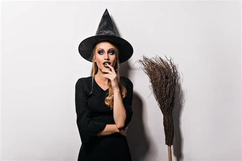Задумчивая женщина волшебник позирует на белой стене чувственная молодая ведьма в черной шляпе