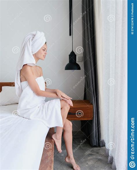 Woman Sitting On Bed After Shower With Wet Hair With Towel On Head In Bedroom And Stroking Body