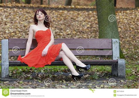 Girl In Elegant Red Dress Sitting On Bench In Autumnal