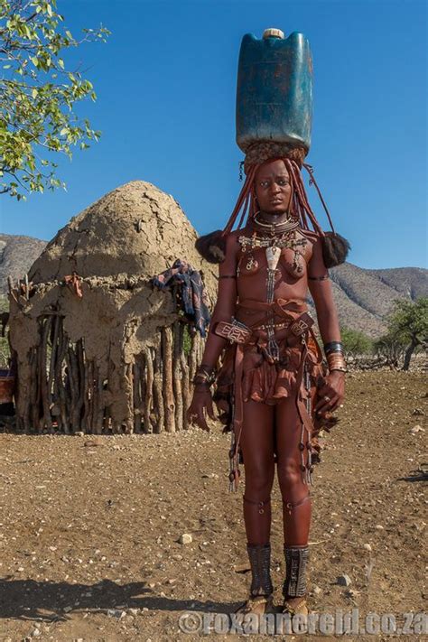 An African Woman In Traditional Garb And Headdress Stands Outside Her Hut With A Bucket On Her Head