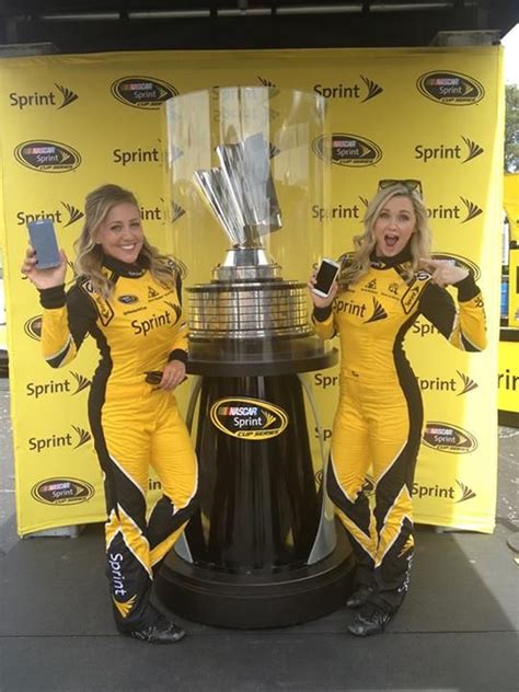 Two Women In Yellow Racing Suits Standing Next To A Large Black Trophy And Posing For The Camera