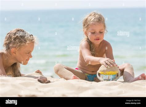 Two Girls On The Beach On A Sunny Day Playing With Sand And A Bucket On A Background Of The Sea