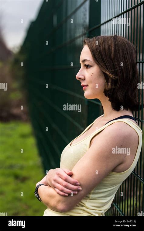 Portait Of A Busty College Girl With Bare Arms Standing Against A Metal Fence In A City Park
