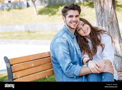 Happy Young Couple In Love Sitting On A Park Bench And Looking At