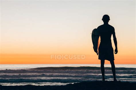 Silhouette Of Man Standing On Beach At Sunset Holding Surfboard — Mid