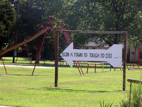 A Sign That Is In The Grass Near Some Swings And Playsets With Trees In