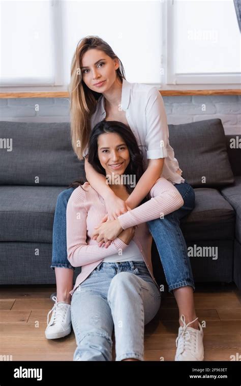 Happy Blonde And Brunette Lesbians Hugging In Living Room Stock Photo