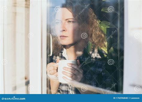 Young Blonde Woman Looking Out Of The Window With A Concerned