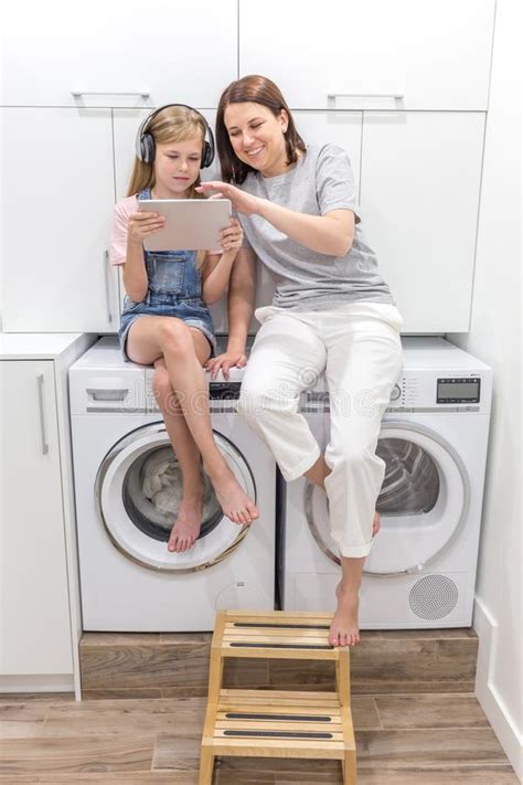 Young Mother And Her Daughter Are Playing With Tablet In Laundry Room
