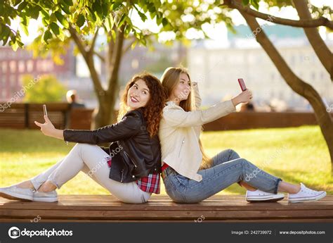 zwei schöne junge frauen machen gleichzeitig selfies einem sonnigen park stockfotografie