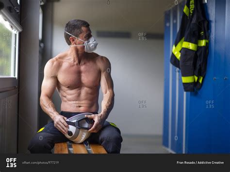 Strong Mature Male Firefighter With Naked Torso Sitting On Bench In Mask And Holding A Helmet