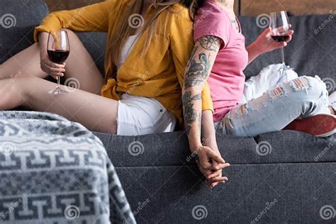 Cropped View Of Two Lesbians Holding Wine Glasses And Holding Hands While Sitting On Sofa In